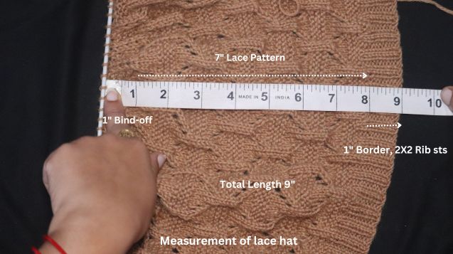 A person using a measuring tape to determine the size of a knitted lace hat laid out on a flat surface.